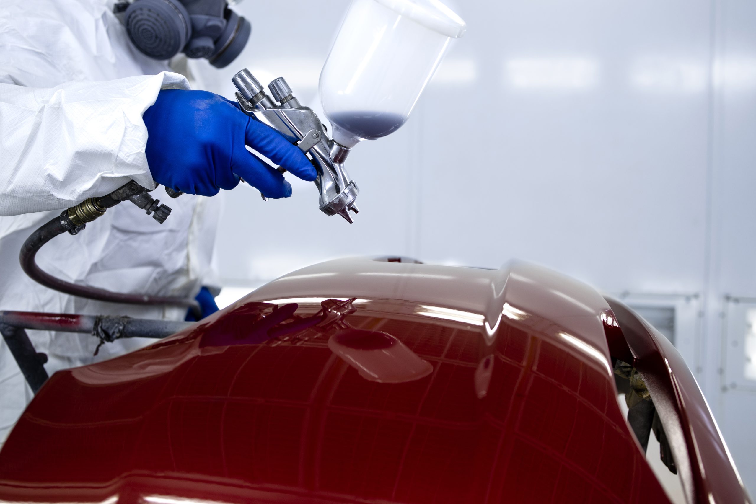 car painter in protective clothes and mask painting automobile bumper with metallic paint and varnish in chamber workshop.